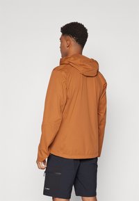 Water-resistant brown jacket with a hood, featuring a smooth texture and a slightly loose fit. Paired with dark shorts and minimal hardware.