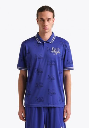 Uomo che indossa una polo blu e pantaloncini abbinati con motivi ripetuti di personaggi dei cartoni animati e la lettera D, in piedi contro uno sfondo bianco.