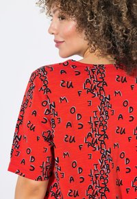 Camiseta roja de manga corta con un patrón de texto negro repetido. La tela parece suave con un acabado ligeramente texturizado.