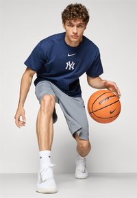 Námornícky modré bavlnené tričko s bielym logom, šedé športové šortky, biele tenisky, drží oranžový basketbal v aktívnej póze.