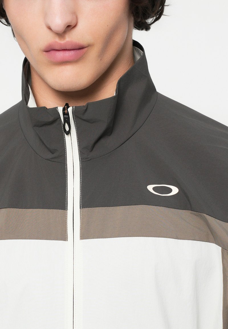 Veste zippée légère gris foncé, blanche et beige avec un col haut. Présente un design de panneaux contrastants et un logo de la marque sur la poitrine.