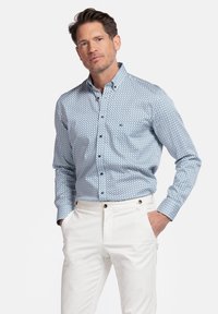Chemise à boutons bleu clair avec un motif géométrique, fabriquée en tissu à la texture lisse ; associée à un pantalon blanc avec des finitions dorées.