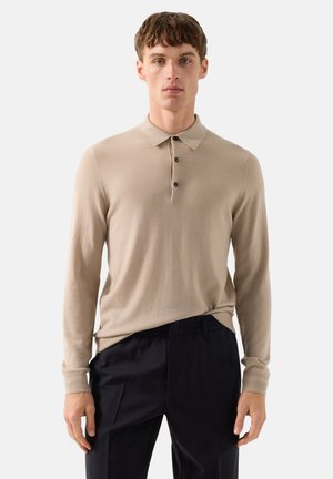 Jonge man die een beige poloshirt met lange mouwen en een zwarte broek draagt, staand tegen een effen lichte achtergrond.