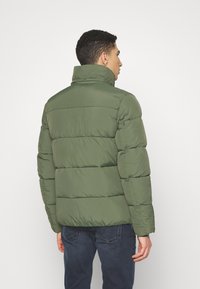 Giacca a vento verde con cuciture orizzontali, colletto alto e texture liscia. Indossata sopra jeans scuri, mostra un design moderno e funzionale.