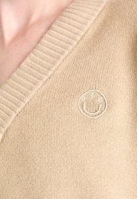 Beżowy sweter z dekoltem w serek z prążkowanymi wykończeniami. Miękka faktura z okrągłym haftowanym logo przypominającym buźkę.