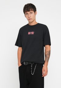 Svart bomull t-skjorte med en rosa AC/DC-logo. T-skjorten har rund hals og avslappet passform, kombinert med svarte bukser og et naglet belte.