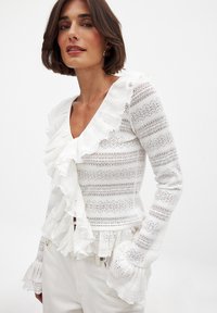 Haut blanc à manches longues avec un décolleté en V, des accents volants, un motif en dentelle et des manches légèrement évasées. Fabriqué en tissu léger et texturé.