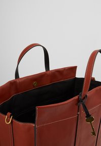 Rote Ledertasche mit schwarzen Akzenten, versehen mit goldenen Hardware-Details und weißer Naht. Offene Oberseite und sichtbare Innentasche mit Reißverschluss.