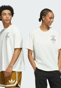 Baltos per didelio dydžio T-shirts su juodu Adidas logotipu ir šriftu. Kairysis modelis dėvi rudus šortus; dešinysis modelis laikosi rankų kišenėse.