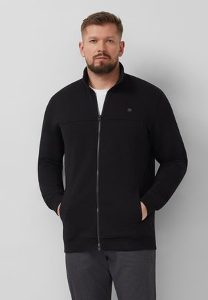 Man met kort haar en baard, gekleed in een zwart jack met ritssluiting, handen in de zakken, staand tegen een effen lichtgrijze achtergrond.