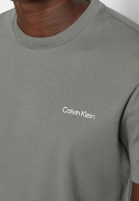 Šedé bavlněné tričko s kulatým výstřihem, které má malou bílou značku "Calvin Klein" na levé straně. Měkká textura a standardní střih.