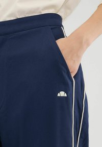 Marineblauwe broek met witte piping, voorzien van een zijzak en een klein logo nabij de tailleband. Gladde stof met een lichte glans.
