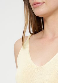 Gros plan sur le bas du visage et le cou d’une femme portant un haut sans manches en maille jaune pâle avec des cheveux bruns lisses.