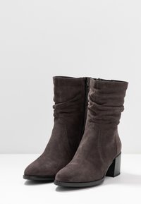 Bottines en daim gris foncé avec un design évasé, bouts pointus, zips sur les côtés et un talon épais. Texture douce avec des détails de couture visibles.