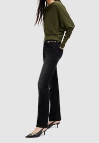 Grüner, drapierter Pullover mit langen Ärmeln, kombiniert mit eng anliegenden schwarzen Jeans und schwarzen Slingback-High Heels mit einer goldenen Schnallenakzent.