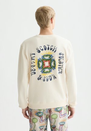 Person trägt einen cremefarbenen Sweatshirt mit buntem Blumendesign und dem Text "Desert Scotch Solstice & South" auf dem Rücken, kombiniert mit gemusterten Hosen.