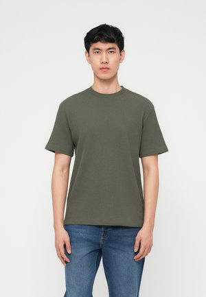 Joven de pie, mirando al frente, con una camisa de manga corta texturizada de color verde oliva y pantalones vaqueros azules, sobre un fondo blanco.