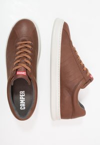 Zapatilla de cuero marrón con parte superior texturizada, suela de goma blanca y cordones marrones. Presenta el logotipo de la marca en la lengüeta y sutil costura lateral.