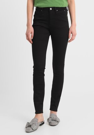 Henkilö, jolla on mustat kapeat farkut, vihreä yläosa ja harmaat ruudulliset slip-on-kengät rusetilla, seisomassa valkoisen taustan edessä.