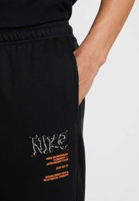 Pantalones de chándal negros de Nike con cinturilla elástica, que presentan letras en blanco y naranja. El diseño incluye bolsillos laterales y un tejido texturizado.