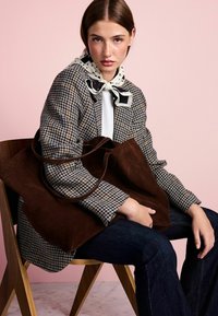 Brown suede tote bag with long handles, worn on a plaid jacket featuring beige and navy tones, paired with dark jeans. Pink background.