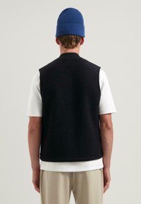 Svart ullväst utan ärmar, med en ribbad krage och texturerad yta. Bärs över en vit t-shirt och krämfärgade byxor, toppad med en blå mössa.
