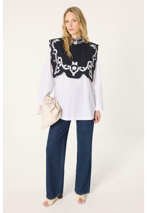 Femme en haut blanc long avec bavette noire décorative, jean bleu à jambes larges, pochette beige froncée et talons ouverts beiges sur fond blanc.