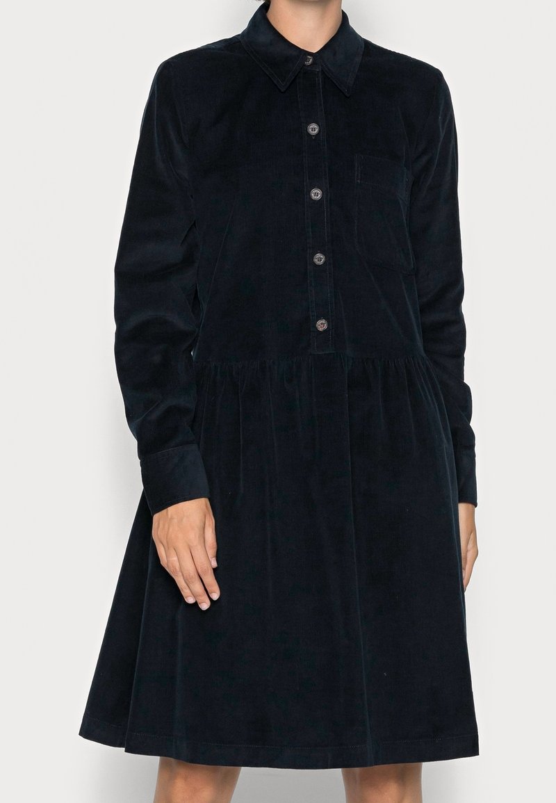 Femme portant une robe noire à manches longues boutonnée avec col et taille froncée, debout devant un fond clair uni.