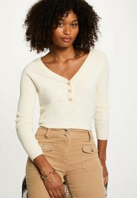 Top bianco a maniche lunghe con righe, scollatura a V e dettaglio con tre bottoni, abbinato a pantaloni beige con tasche e accenti metallici.
