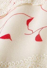 Tessuto bianco con motivi astratti rossi, con pizzo lungo il bordo. La texture appare liscia con una leggera lucentezza.