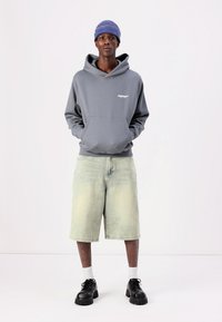 Grijze hoodie met voorzak en logo, gecombineerd met lichtgroene denim shorts. Zwarte chunky schoenen met witte sokken. Blauwe gebreide beanie.