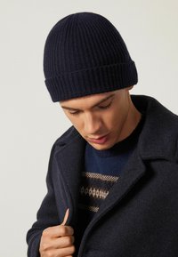 Marinblå stickad mössa med ribbad textur, åtsittande design och uppvikt kant. Buren med en mörk jacka och en mönstrad tröja under.