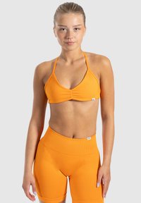 Sostén deportivo naranja con tirantes ajustables y un detalle fruncido en el frente, combinado con pantalones cortos de ciclismo naranjas de cintura alta, ambos hechos de tela elástica.