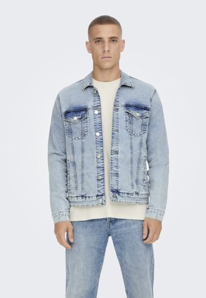 Light blue denim jacket with a classic design, featuring button-up front, two chest pockets, and contrast stitching. Worn over a beige shirt.