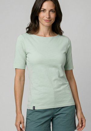 Frau mit schulterlangem dunklem Haar, die ein hellgrünes Kurzarmshirt und türkisfarbene Shorts trägt, vor einem schlichten Hintergrund stehend.