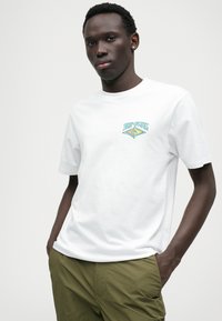 Camiseta branca de algodão com um logótipo "Rip Curl" multicolorido do lado esquerdo, combinada com calções verdes-oliva. Design simples.