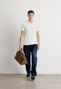 Hvit polo skjorte, mørkeblå jeans og svarte sko. Bærer en brun, teksturert veske med flere lommer. Ensfarget bakgrunn.