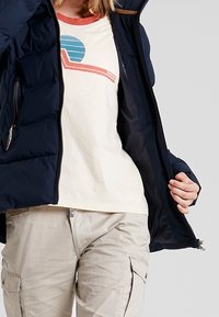 Tmavě modrá péřová bunda s kapsami na zip, nošená přes krémové tričko s grafikou v tyrkysové a červené barvě. Světle béžové kalhoty typu cargo.