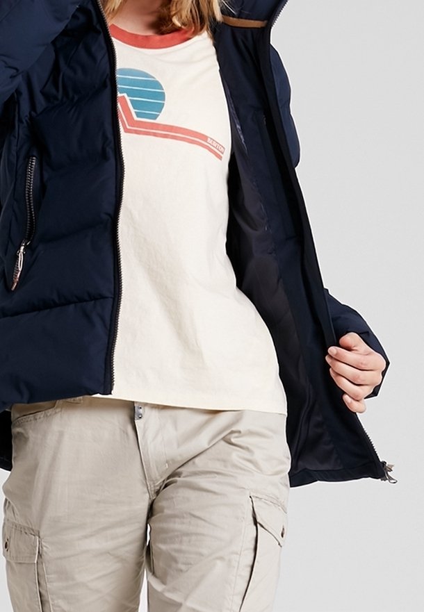 Tmavě modrá péřová bunda s kapsami na zip, nošená přes krémové tričko s grafikou v tyrkysové a červené barvě. Světle béžové kalhoty typu cargo.