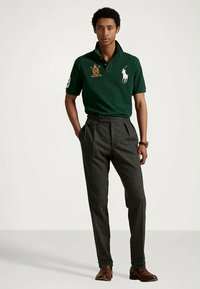 Zielona koszulka polo z krótkim rękawem z haftowanym emblematem i logo konia, zestawiona z szarymi spodniami w kant i brązowymi butami.