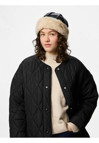 Černá prošívaná bunda s patentkami, nošena přes krémový svetr. Hnědá čepice s kožíškem dotváří celý outfit. Viditelné kudrnaté vlasy.