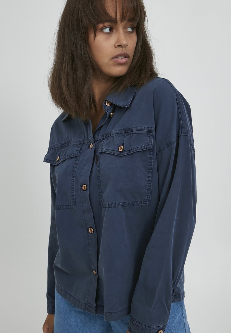 Chemise boutonnée bleu marine avec deux poches poitrine, boutons cuivrés et coupe décontractée. Présente un col classique et une texture douce.