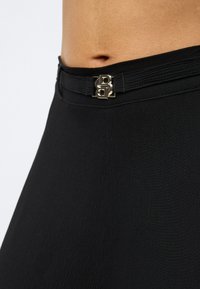 Melnas bikses ar augsto vidukli, teksturētu audumu, plakanu jostasvietu un zelta logotipa sprādzi priekšpusē aizdarīšanai.