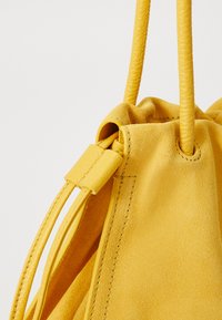 Sac en daim jaune avec une fermeture à cordon, corps texturé et accents en cuir lisse. Dispose de deux anses tressées.