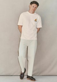 White cotton t-shirt with orange and green graphic logo, paired with light-colored jeans and dark brown shoes with tan laces and chunky soles.