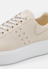 Baskets en cuir beige avec lacets blancs, œillets dorés, motif latéral perforé et semelle épaisse en caoutchouc blanc sur un fond uni.