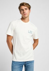 Camiseta de algodón blanca con cuello redondo, mangas cortas y un logo azul de "LEE" en el lado izquierdo del pecho. Llevada con unos pantalones vaqueros azules.