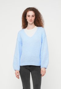 Νέα γυναίκα με σγουρά κόκκινα μαλλιά, που φοράει ανοιχτό μπλε πουλόβερ με V λαιμόκοψη και μαύρο τζιν, στέκεται μπροστά σε απλό λευκό φόντο.