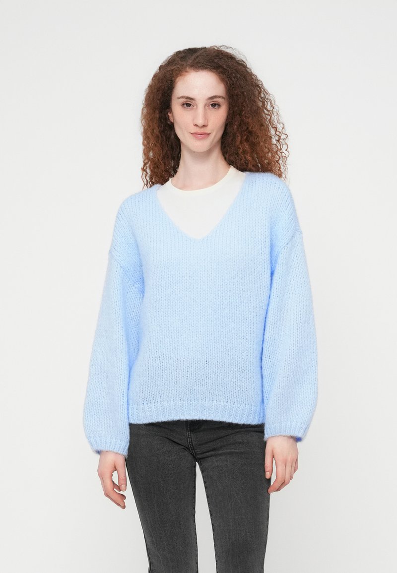 Νέα γυναίκα με σγουρά κόκκινα μαλλιά, που φοράει ανοιχτό μπλε πουλόβερ με V λαιμόκοψη και μαύρο τζιν, στέκεται μπροστά σε απλό λευκό φόντο.