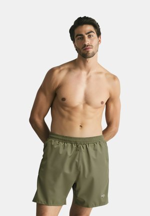 Uomo con capelli scuri che indossa pantaloncini da bagno verde oliva, in piedi contro uno sfondo semplice, con le mani dietro la schiena, che guarda verso la fotocamera.